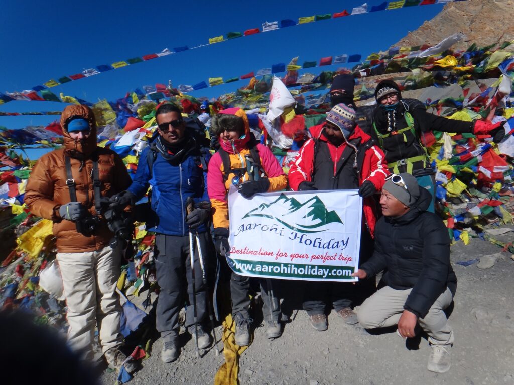 Group of trekkers from Aarohi Holiday at Thorong La Pass in the Annapurna region, surrounded by colorful prayer flags.