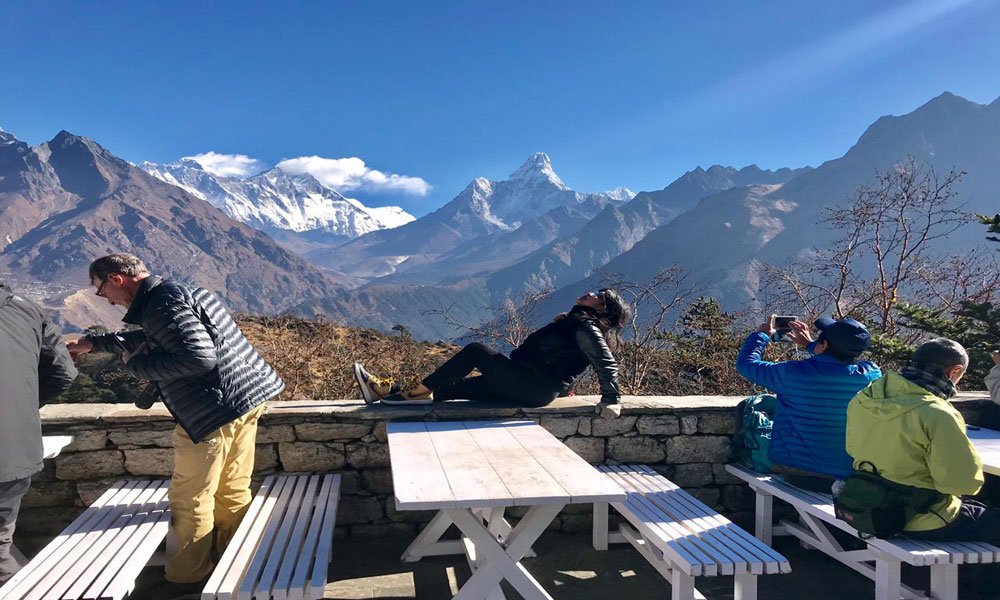 A group of travelers enjoying a panoramic view of snow-capped Himalayan peaks, including Mount Everest and Ama Dablam, from an outdoor stone patio with white picnic tables under a clear blue sk