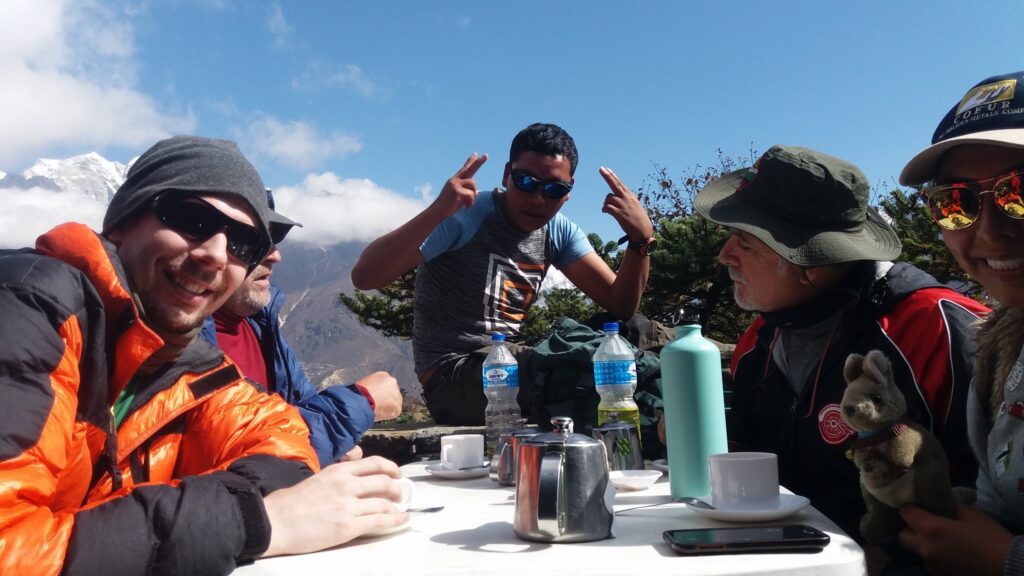 Trekkers enjoying tea and sunshine at Everest View with Himalayan peaks in the background.